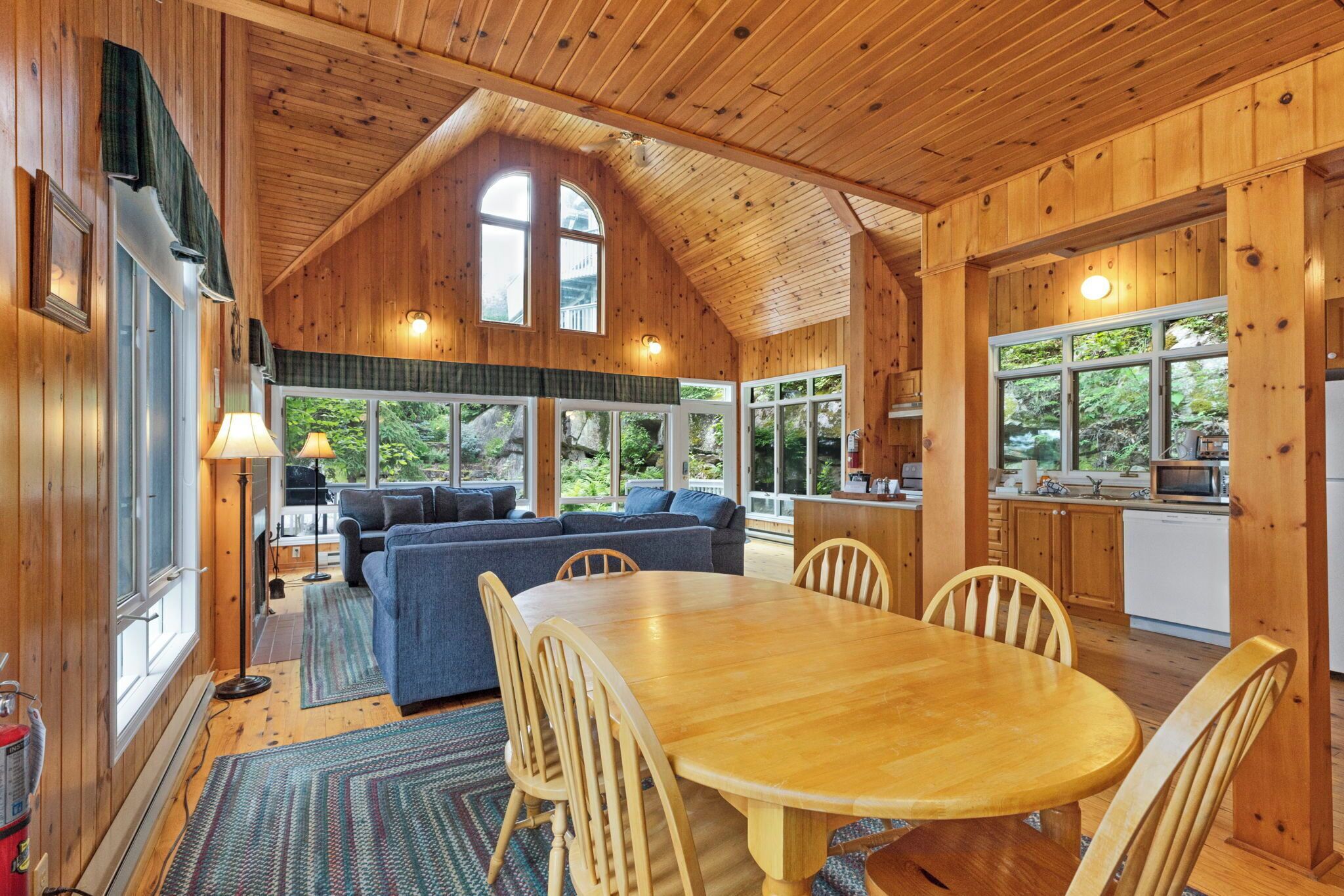A wooden cabin interior with a dining area, sofa, kitchen, and large windows.