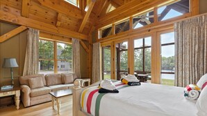 Ein Schlafzimmer einer Holzhütte mit einem Bett, einem Schlafsofa, einem Schreibtisch und großen Fenstern mit Blick ins Freie.