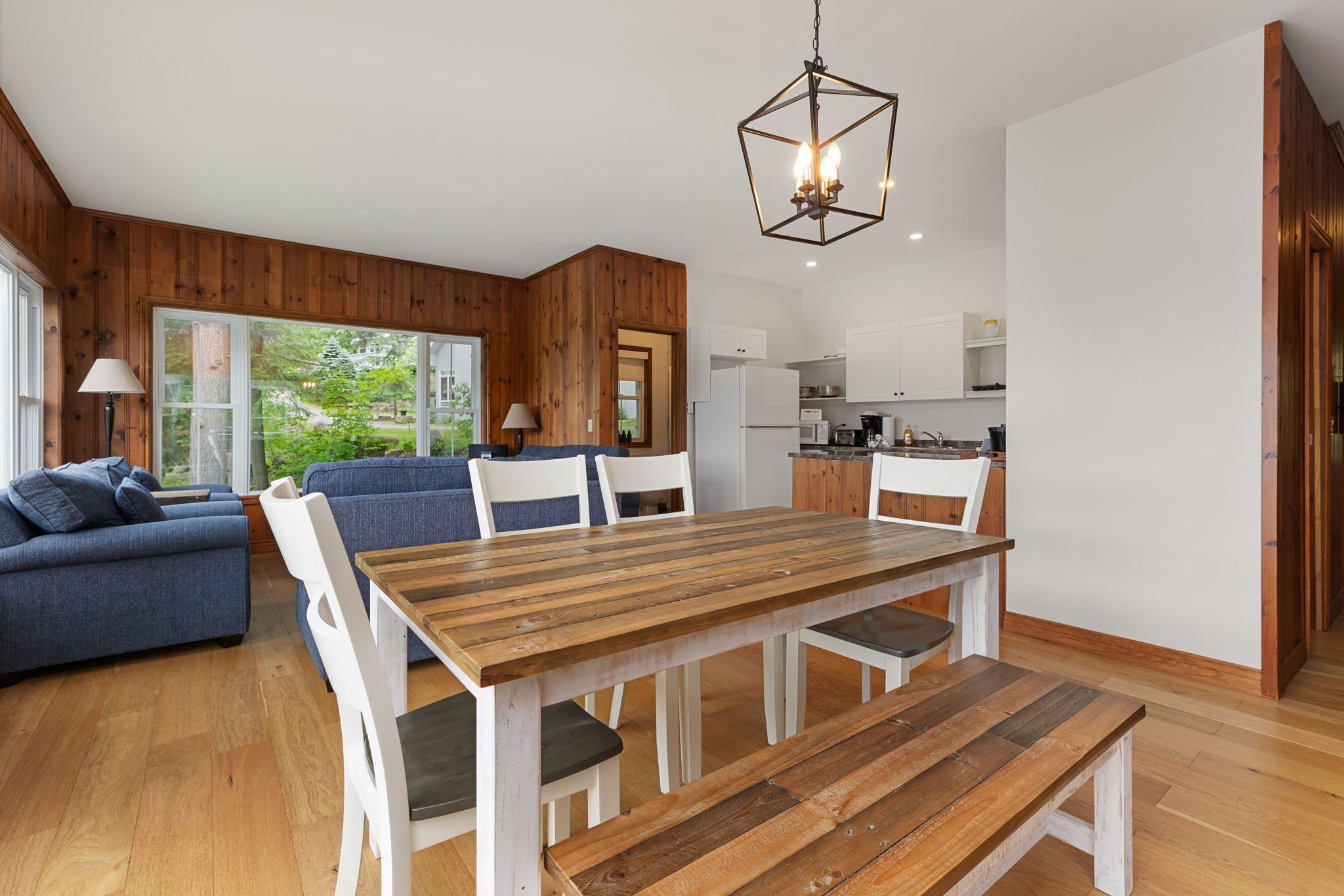 A modern living room with a wooden dining table, blue sofa, and white chairs. A kitchen is visible in the background with white cabinets and a refrigerator.
