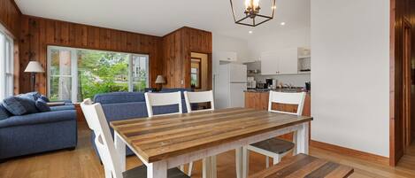 A modern living room with a wooden dining table, blue sofa, and white chairs. A kitchen is visible in the background with white cabinets and a refrigerator.
