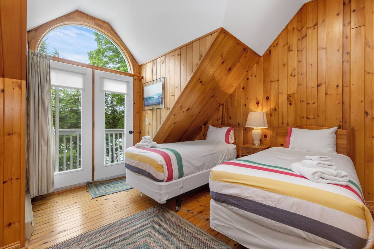 A bedroom with two beds, wooden walls, a large window, and a door leading to a balcony.
