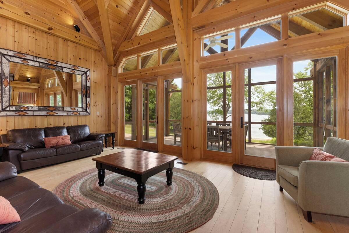 A spacious living room with a wooden ceiling, large windows, a brown leather sofa, a gray armchair, and a wooden coffee table.