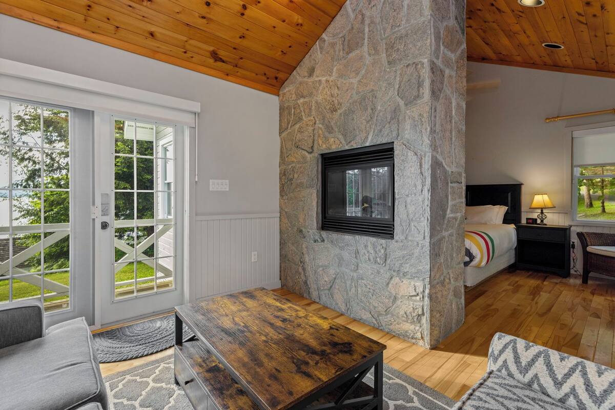 A cozy room with a stone fireplace, a bed with a colorful blanket, a wooden table, and a view of a green landscape through large windows.