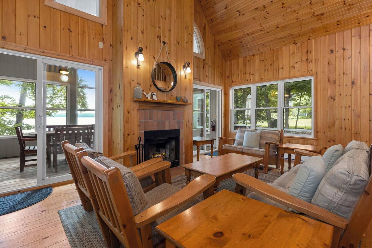 A cozy wooden cabin interior with a fireplace, wooden furniture, and large windows offering views of the outdoors.