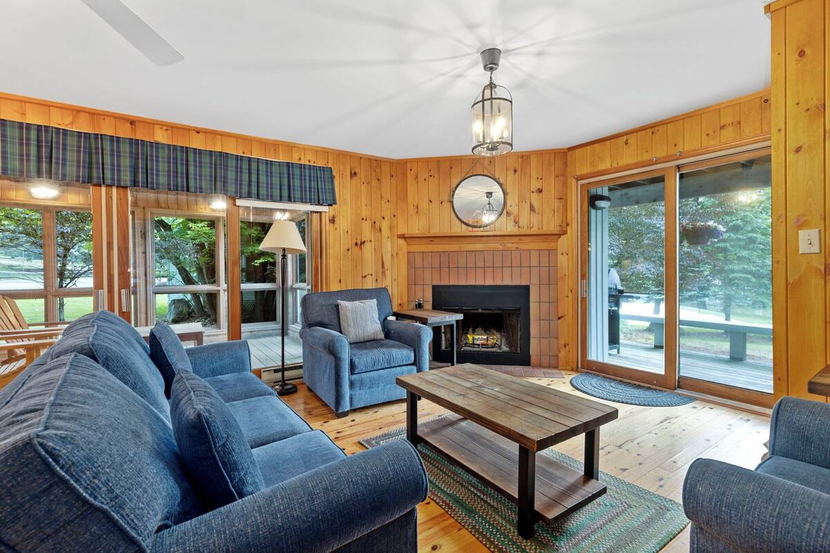 A cozy living room with a fireplace, wooden walls, and large windows.
