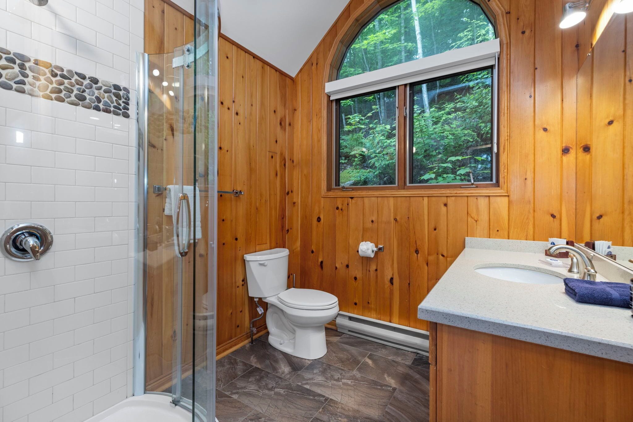 A bathroom with a glass shower enclosure, a toilet, and a sink with a window above.
