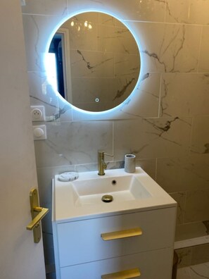 Une salle de bain avec un lavabo blanc, un robinet doré, un miroir rond avec un éclairage intégré et des murs à motifs de marbre.
