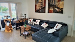 A modern living room with a black leather sofa, decorative pillows, a small wooden table with a vase, and a view of a cityscape through large windows.