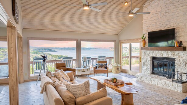 A spacious living room with a stone fireplace, a large TV mounted above it, and a view of a lake through large windows. - Lake Home with Private Pool, Games, Views! 100 yds to Marina w boat launch! (Strawn)