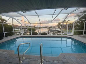 A pool with clear water, surrounded by a glass enclosure, offering a view of a lake and distant buildings. - Lovely Home with Canal View, Heated Pool, Boatlift, Stunning Sunsets (Cape Coral)