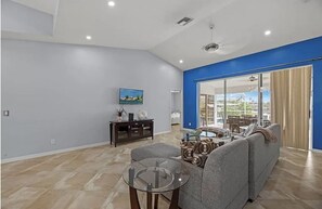 A spacious living room with a grey sofa, a glass coffee table, and a television on a wall. - Lovely Home with Canal View, Heated Pool, Boatlift, Stunning Sunsets (Cape Coral)