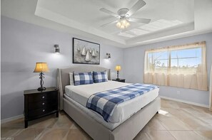 A bedroom with a large bed, two bedside tables, a ceiling fan, and a window with curtains. - Lovely Home with Canal View, Heated Pool, Boatlift, Stunning Sunsets (Cape Coral)