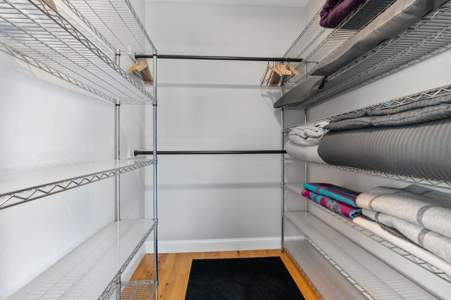 A walk-in closet with metal shelving, a central rod for hanging clothes, and a black rug on the floor.