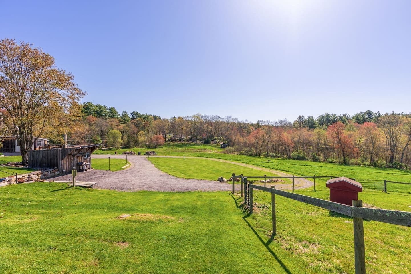A rural landscape with a dirt path leading to a wooden structure, surrounded by green fields and autumn-colored trees.