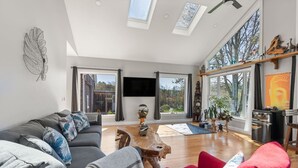 Amplia sala de estar con un sofá gris, un sillón rojo y una mesa de centro de madera. Grandes ventanales ofrecen vistas al exterior.