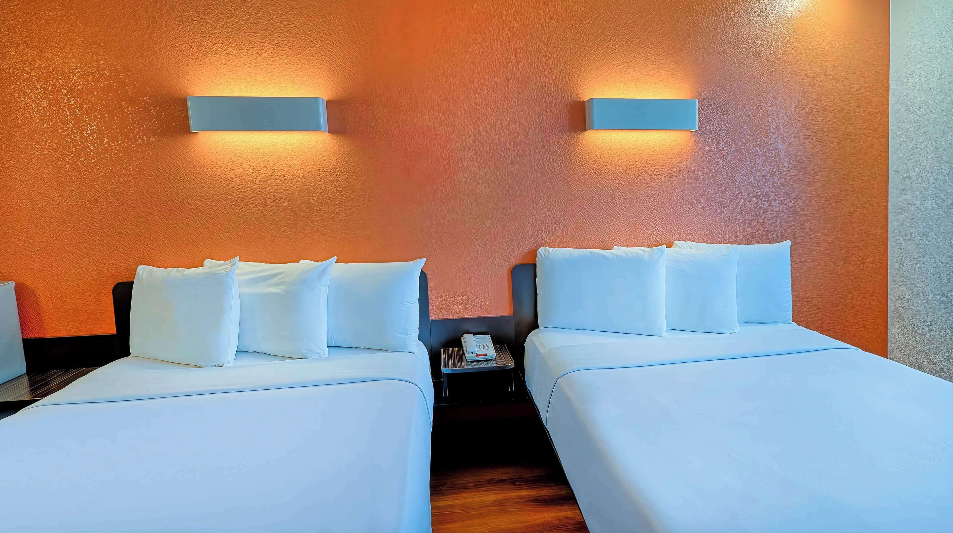 Habitación de hotel con dos camas, ropa de cama blanca, una mesita con un teléfono y luces fijadas a la pared.