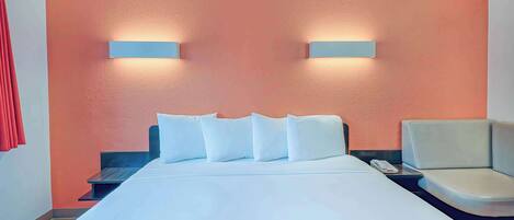 Una habitación de hotel con una cama, almohadas blancas, una mesita de noche, un teléfono y una silla.