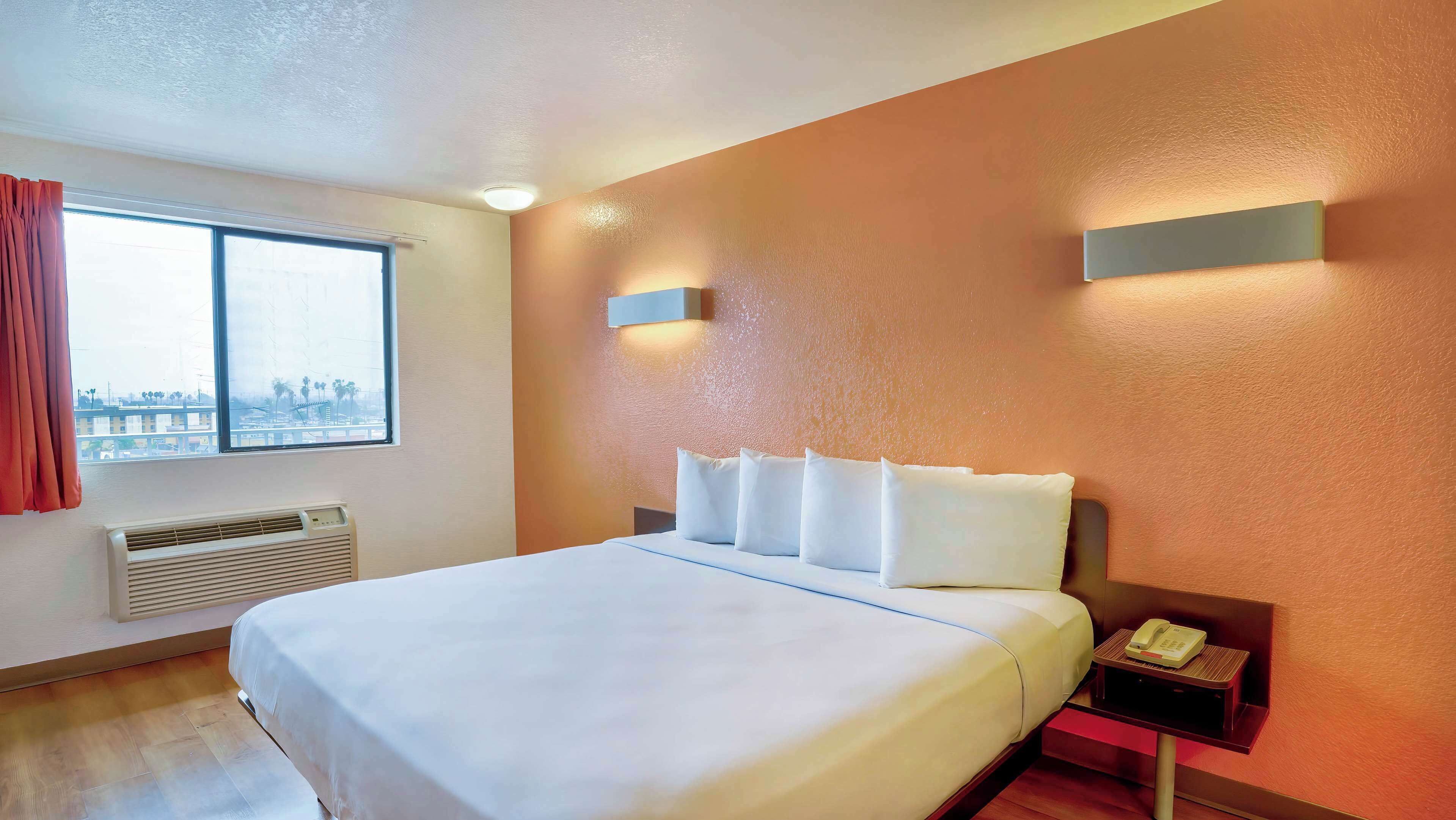 A hotel room with a large bed, a window with curtains, a wall-mounted phone, and a nightstand with a lamp.