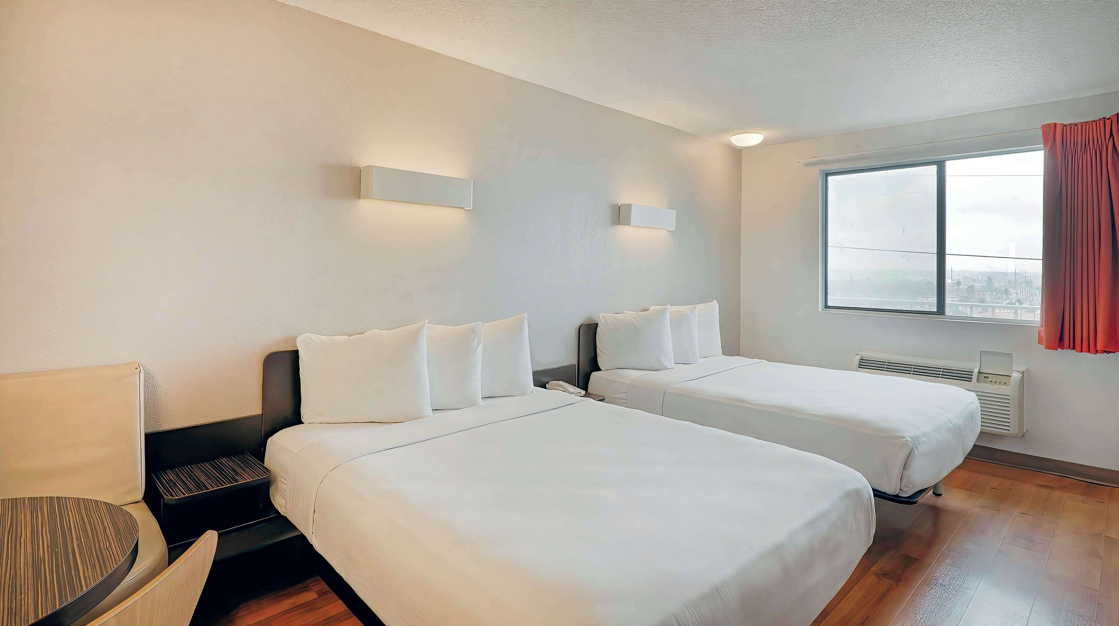 Een hotelkamer met twee bedden, een bureau, een stoel, een raam met gordijnen en uitzicht op de stad.
