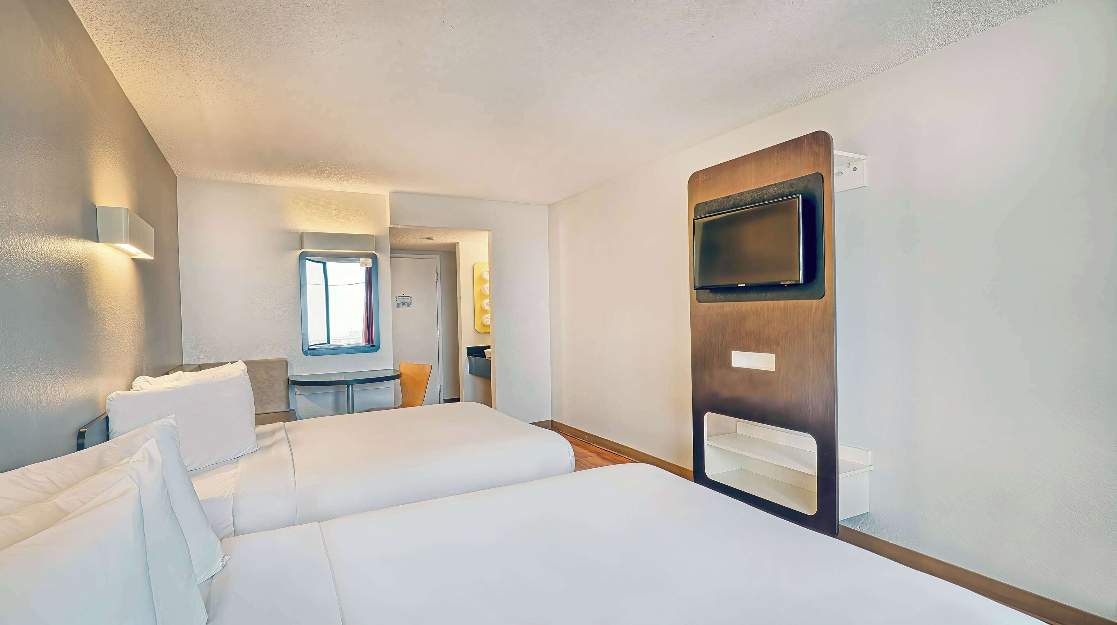 Een hotelkamer met een bed, een klein tafeltje, een stoel, een aan de muur bevestigde televisie en een raam.