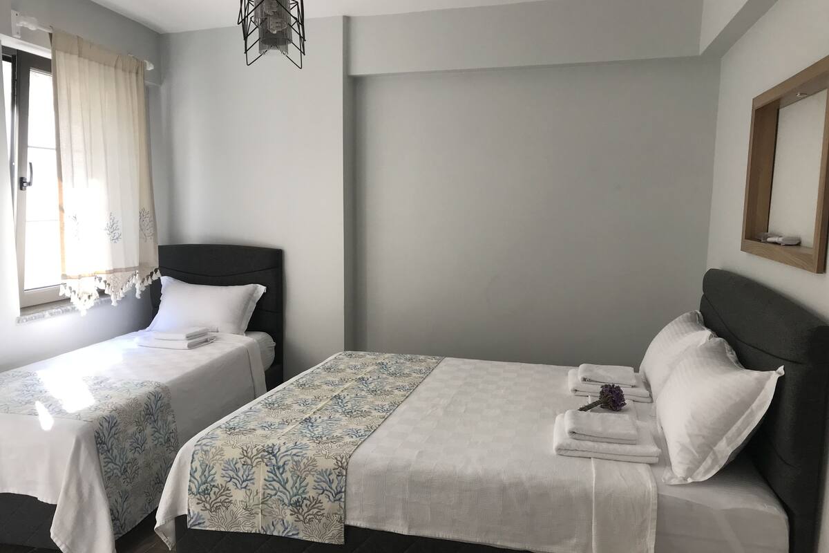 A hotel room with two beds, each with a dark headboard and white bedding. There is a window with curtains, a wall-mounted light fixture, and a mirror above the right bed.