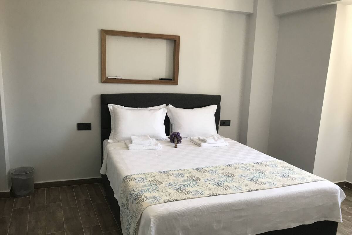 A neatly made bed with white linens and a floral-patterned bedspread, a dark headboard, and a small wooden-framed mirror above it.