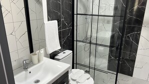 A modern bathroom with a white sink, dark marble tiles, a glass-enclosed shower, and a white toilet. - Demirtaş Pansiyon (Bozcaada)