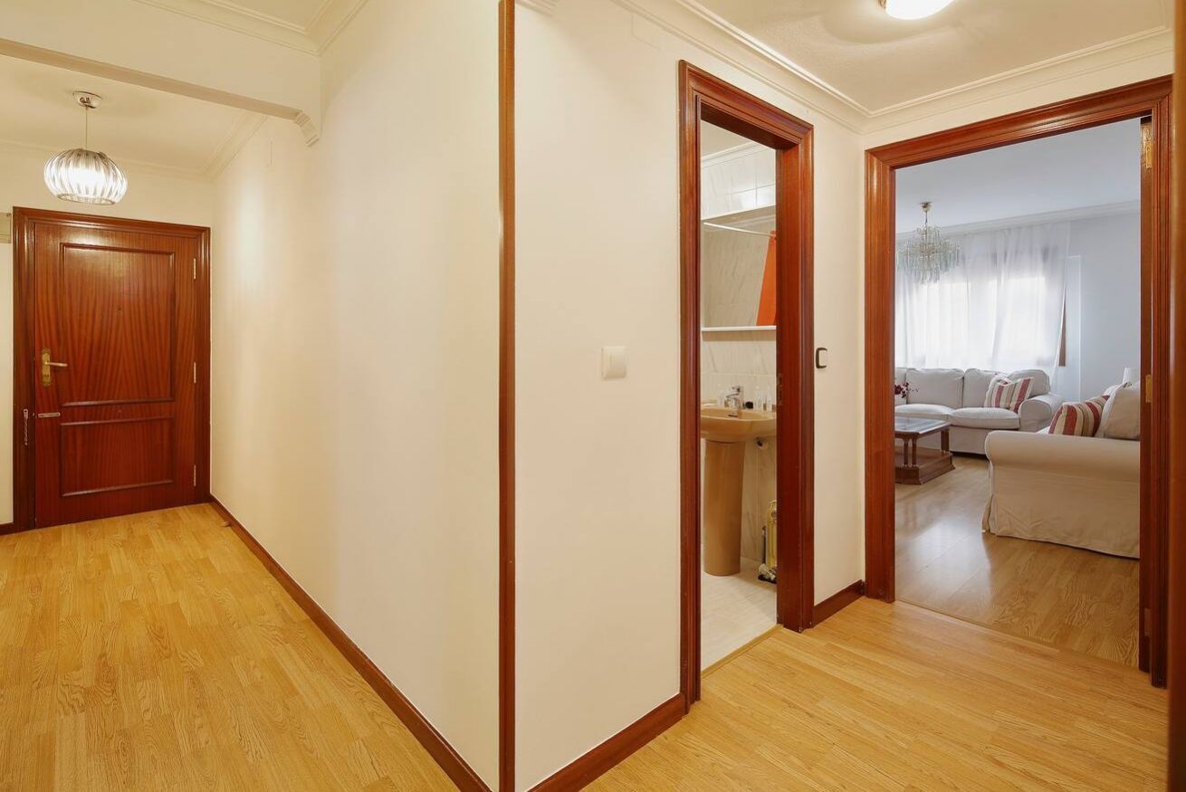 Ein Flur mit Holzboden, einer Holztür und einem Spiegel, der ein Wohnzimmer mit Sofa und Fenster wiederspiegelt.