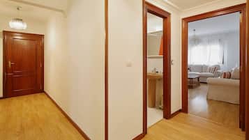 Un pasillo con suelo de madera, una puerta de madera y un espejo que refleja una sala de estar con un sofá y una ventana.