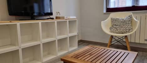 Una sala de estar con un televisor de pantalla plana sobre un soporte de madera, una silla blanca con un cojín estampado y una mesa de centro de madera.