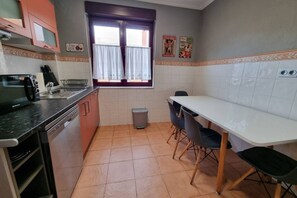 Eine kompakte Küche mit Spülmaschine, Spüle, einem Fenster mit Vorhängen, einem Esstisch mit Stühlen und einem Mülleimer.