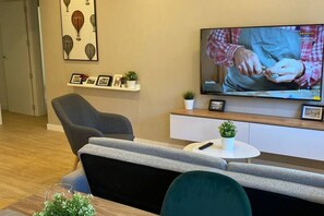 Een moderne woonkamer met een bank, een televisie, een bijzettafeltje met een plant en een wandplank met ingekaderde foto's.