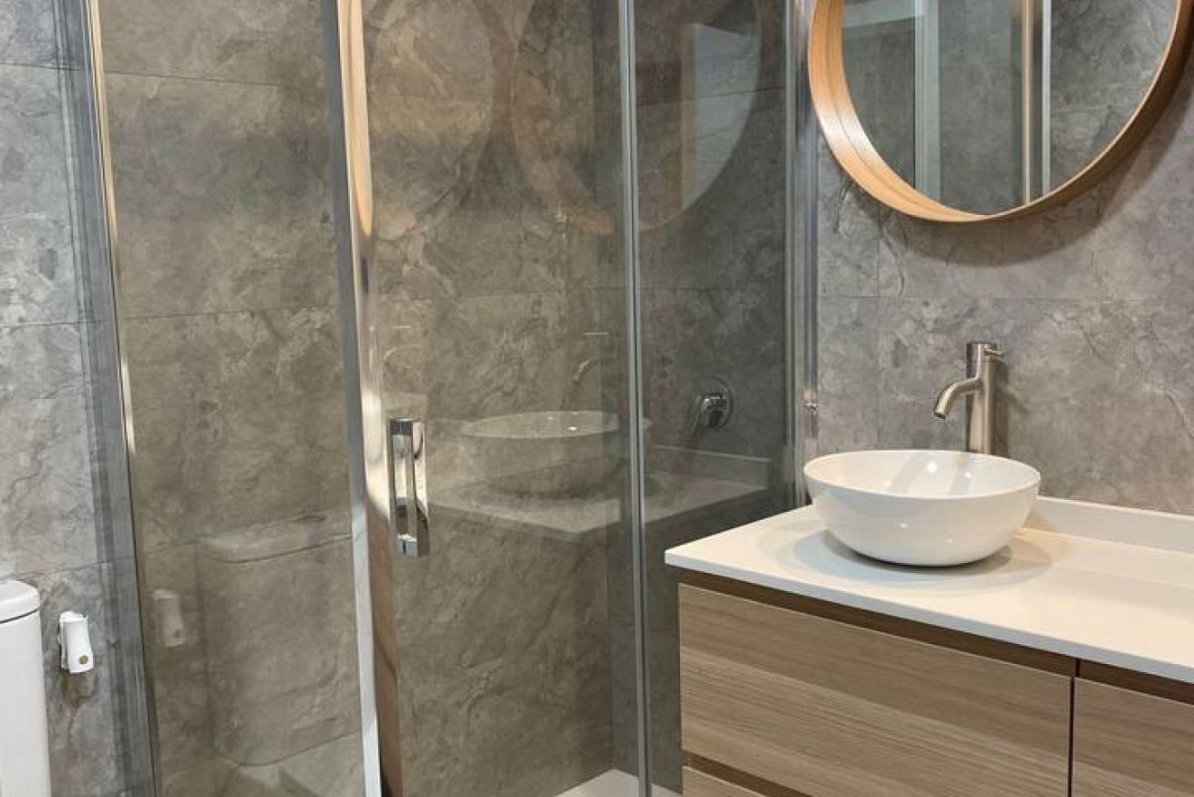 Un baño moderno con cabina de ducha de vidrio, lavamanos blanco con lavabo redondo y tocador de madera.