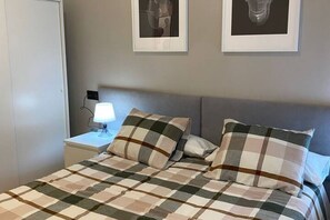 Een bed met een geruite sprei en kussens, een nachtkastje met een lamp, en twee ingelijste foto's aan de muur.