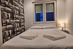 Habitación de hotel con una cama grande, dos almohadas, una ventana con persianas y un mural de paisaje urbano en la pared, además de una mesita de noche con una lámpara.