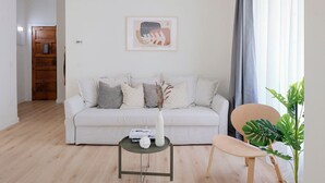 Ein modernes Wohnzimmer mit einem weißen Sofa, einem kleinen runden Tisch mit einer Vase und einem Buch, einem Holzstuhl und einem großen Fenster mit Vorhängen.