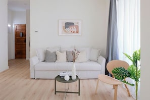 Ein modernes Wohnzimmer mit einem weißen Sofa, einem kleinen runden Tisch mit einer Vase und einem Buch, einem Holzstuhl und einem großen Fenster mit Vorhängen.
