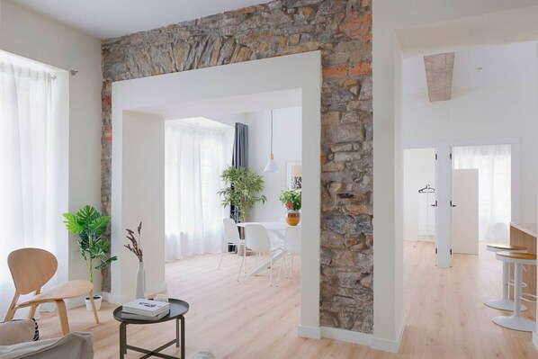 A modern living room with a stone accent wall, a dining area with white furniture, and a kitchen area with bar stools.