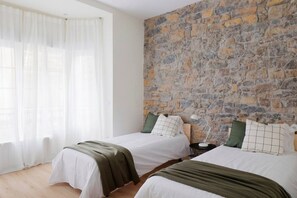 A room with two beds, a stone wall, and a window with sheer curtains.