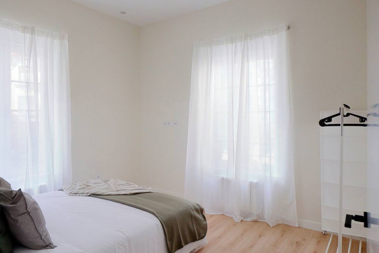 A bedroom with a bed, a bedside table, a hanging coat rack, and a window with sheer curtains.