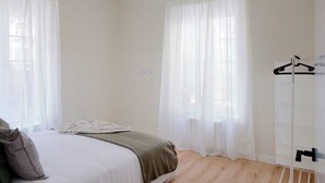 Ein Schlafzimmer mit einem Bett, einem Nachttisch, einem Kleiderhaken und einem Fenster mit durchsichtigen Vorhängen.