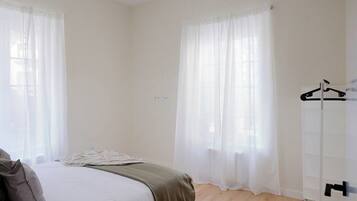 A bedroom with a bed, a bedside table, a hanging coat rack, and a window with sheer curtains.