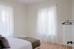 Ein Schlafzimmer mit einem Bett, einem Nachttisch, einem Kleiderhaken und einem Fenster mit durchsichtigen Vorhängen.