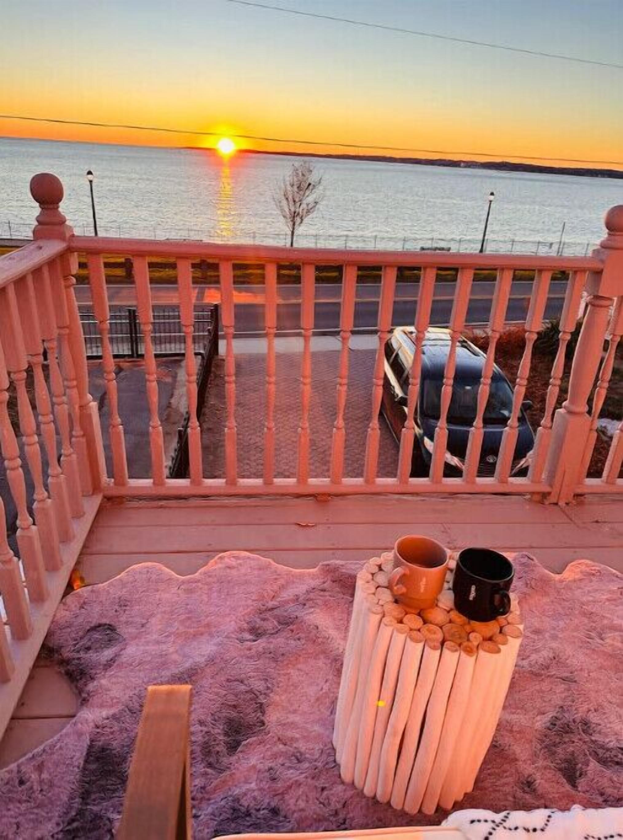 A balcony with a view of the sunset over water, a car parked in the background, and a small table with a cup and a mug.