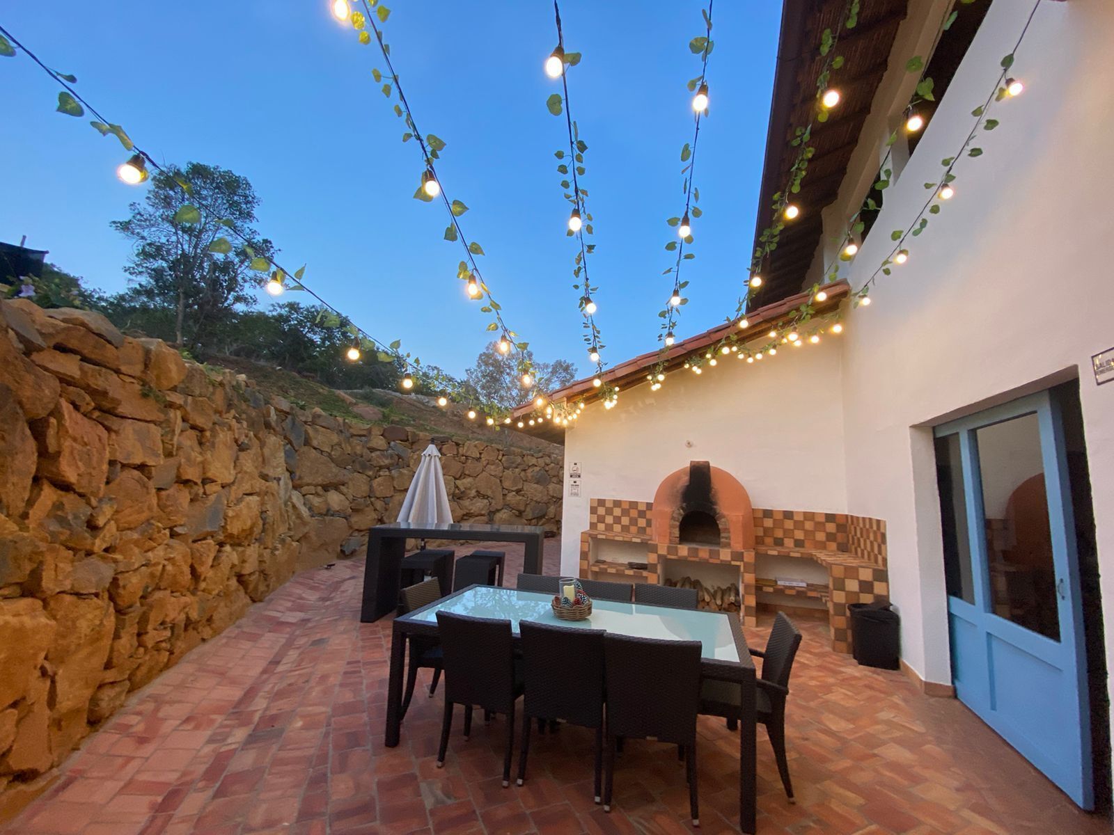 Un patio al aire libre con paredes de piedra, una mesa de vidrio, sillas y un horno para pizza.
