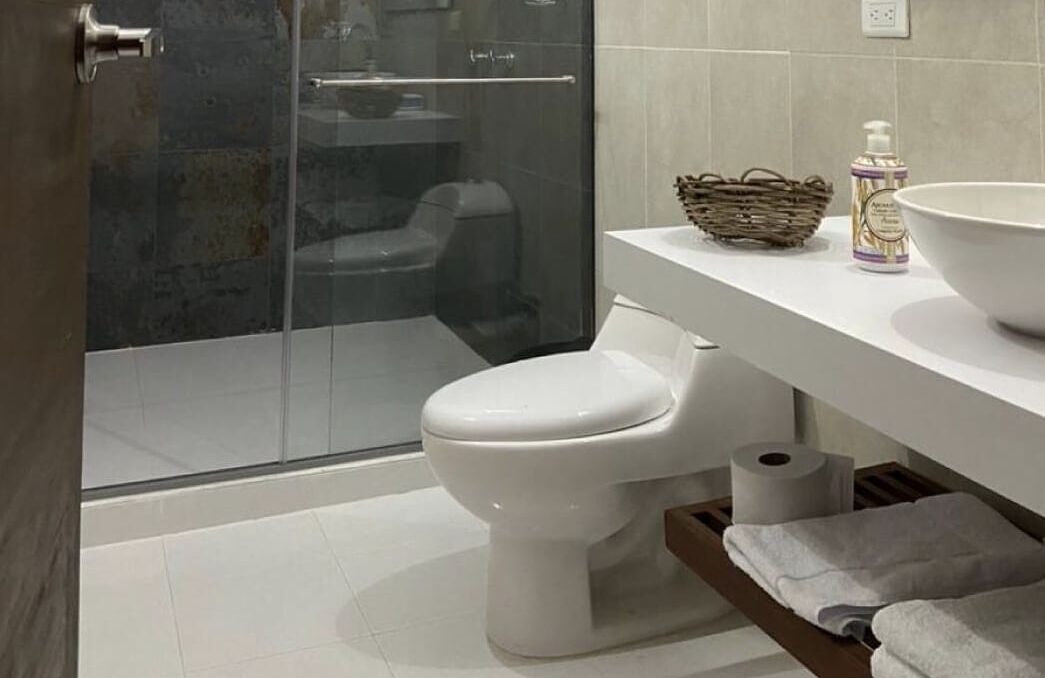 A modern bathroom with a glass shower enclosure, a white toilet, a white sink with a large bowl, a wicker basket, and a bottle of hand soap.
