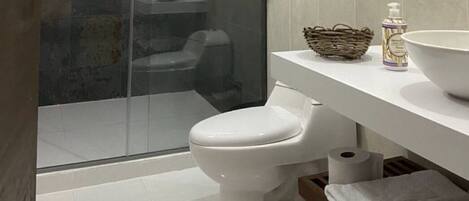 A modern bathroom with a glass shower enclosure, a white toilet, a white sink with a large bowl, a wicker basket, and a bottle of hand soap.