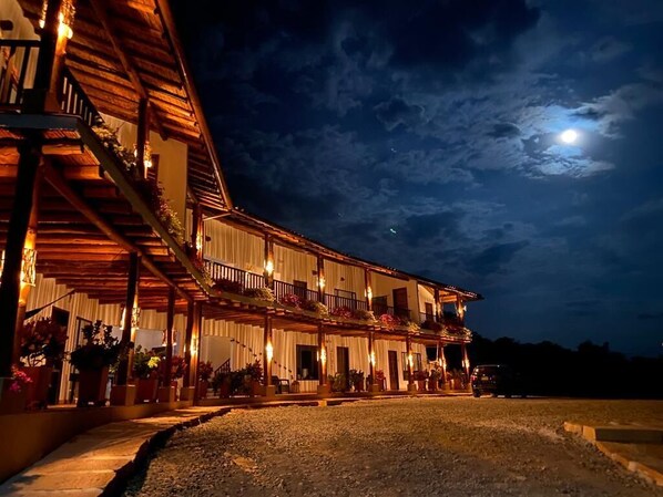 Un bâtiment à deux étages doté d’un balcon et d’un espace extérieur aménagé pour s’asseoir, sous un ciel nocturne où l’on peut voir la lune.