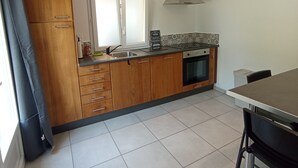 A compact kitchen with wooden cabinets, a built-in oven, and a window. - Le Penty Gîte (Plestin-les-Grèves)