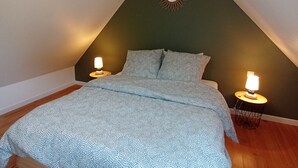A bedroom with a sloped ceiling, a bed with patterned bedding, two bedside lamps, and a decorative sunburst mirror on the wall. - Le Penty Gîte (Plestin-les-Grèves)